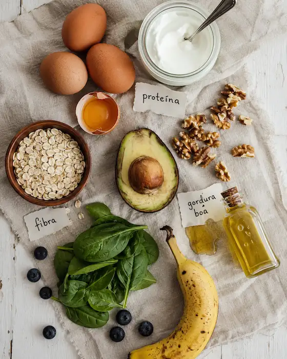 Flat lay de ingredientes: ovos, iogurte, aveia, abacate, espinafres, nozes e azeite — proteína, fibra e gordura boa